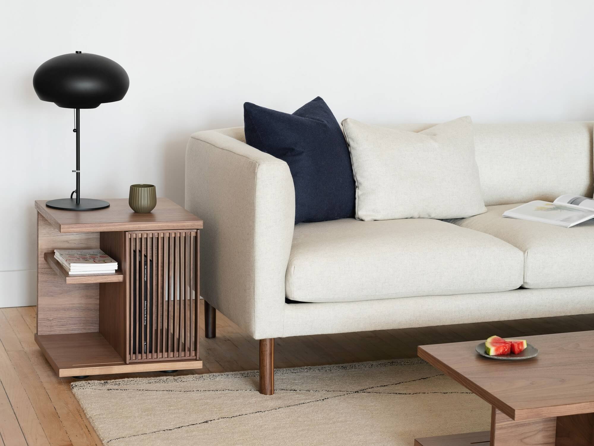 The Plank modern end table in walnut with books, a lamp, and a cup on it sits beside a white sofa and an area rug